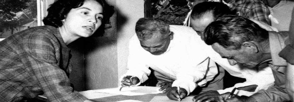 A black and white image of a woman speaking as three men are writing on paper. 